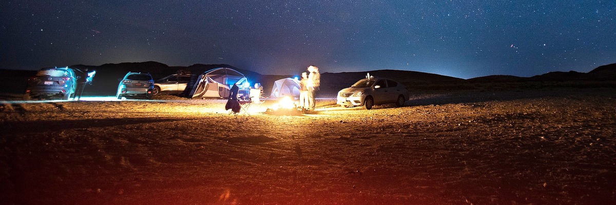 A 1200×400 attention crop of the desert photo with the cars, tents, campfire, and people fully in frame.