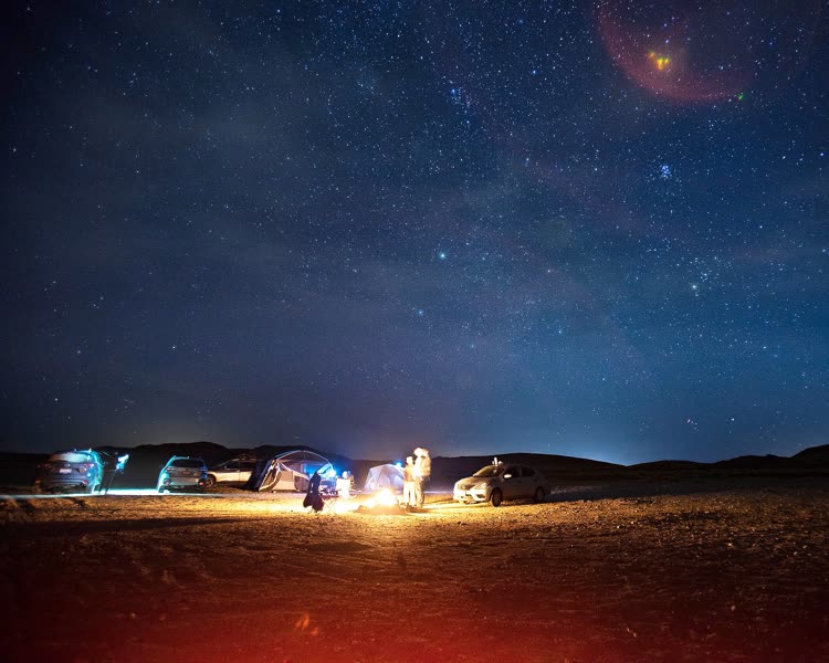 A night photo of a desert campsite with cars, tents, and a campfire in the lower third, under a starry sky.